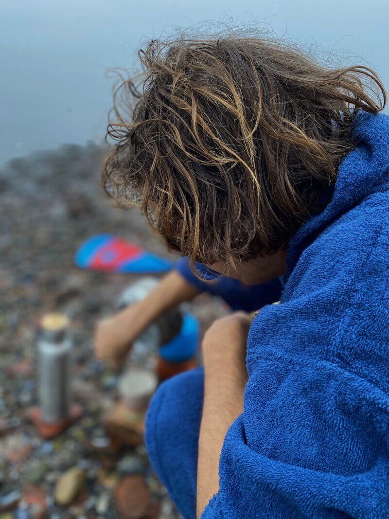 Dreng med krøllet brunt hår der sidder ned med en SURFMORE badeponcho af bomuld i blå på